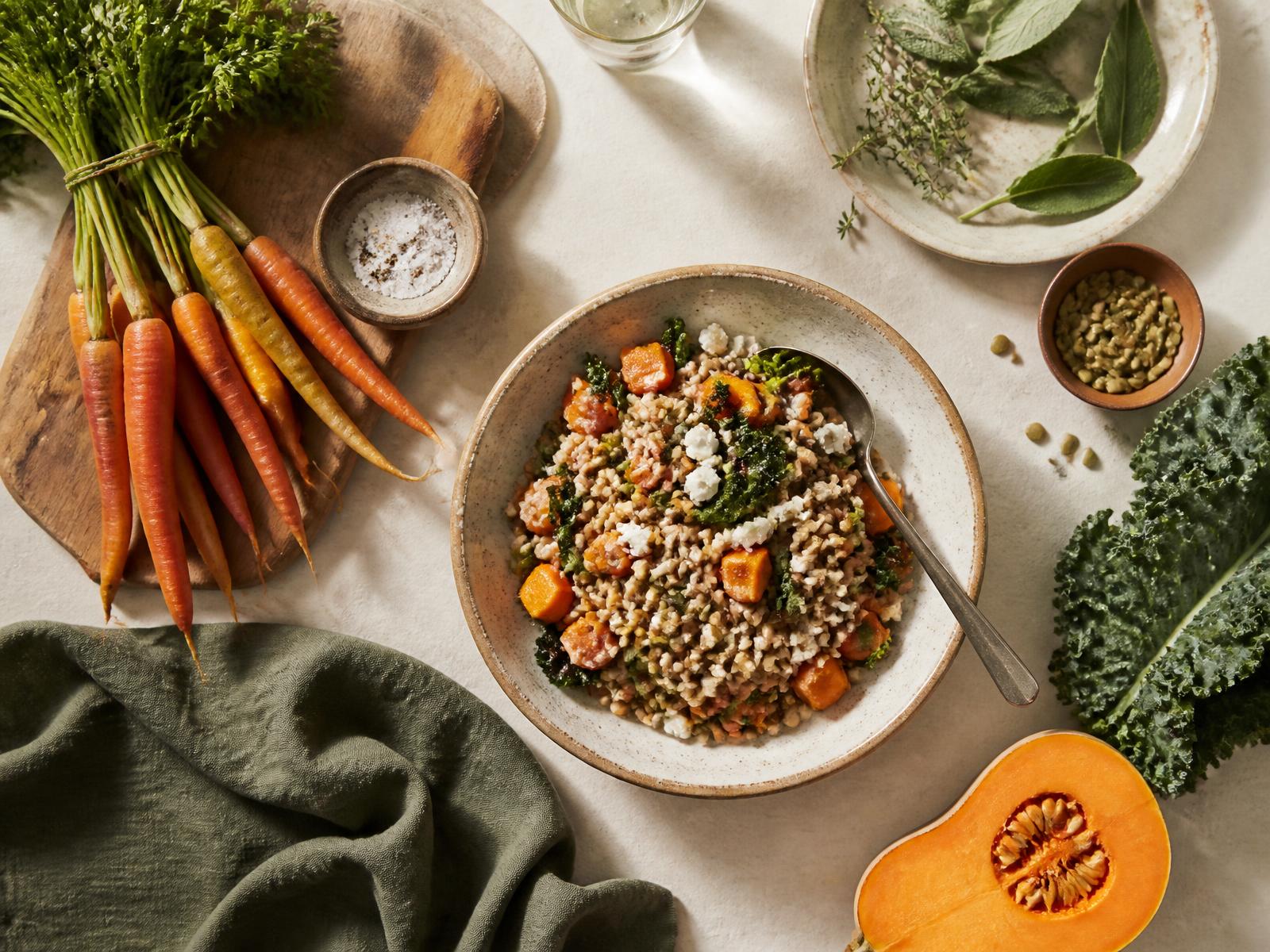 A kitchen table with fresh vegetables, herbs and a grain bowl